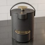 Modern ribbed matte black 2-liter hot water dispenser with gold top button, carrying handle, and gold drip tray on a kitchen countertop.