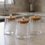 Three hammered glass kitchen storage jars with wooden wooden lids and crystal handles on a white marble countertop