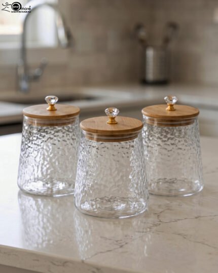 Three hammered glass kitchen storage jars with wooden wooden lids and crystal handles on a white marble countertop