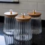 Three ribbed glass storage jars with natural wooden lids topped with clear crystal diamond knobs on a kitchen countertop.