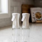 2 Pcs clear glass oil sprayer bottle set with ergonomic white trigger nozzles on a kitchen countertop