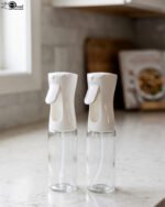 2 Pcs clear glass oil sprayer bottle set with ergonomic white trigger nozzles on a kitchen countertop