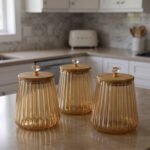 Three ribbed amber glass storage canisters with wooden lids and crystal knobs sitting on a marble kitchen countertop.