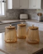 Three ribbed amber glass storage canisters with wooden lids and crystal knobs sitting on a marble kitchen countertop.