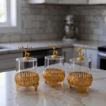 Three clear glass storage jars resting on ornate gold filigree metal stands, each topped with a decorative gold deer figure lid on a white marble kitchen countertop.