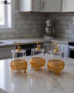 Three clear glass storage jars resting on ornate gold filigree metal stands, each topped with a decorative gold deer figure lid on a white marble kitchen countertop.