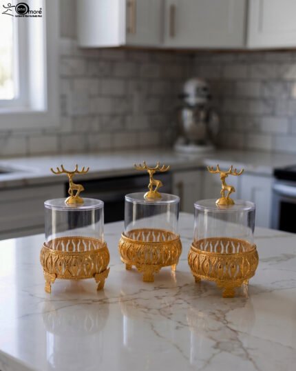 Three clear glass storage jars resting on ornate gold filigree metal stands, each topped with a decorative gold deer figure lid on a white marble kitchen countertop.