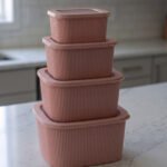 Four dusty rose ribbed plastic storage boxes with lids stacked vertically by size on a white marble kitchen countertop