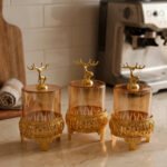Three amber glass storage jars with gold deer lid handles on intricate gold filigree stands set on a kitchen marble countertop next to an espresso machine.