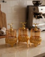 Three amber glass storage jars with gold deer lid handles on intricate gold filigree stands set on a kitchen marble countertop next to an espresso machine.