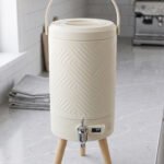 Modern 5L cream beverage dispenser with wooden legs, digital temperature display, and chrome spigot sitting on a marble kitchen countertop.