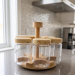 Six clear glass spice jars with smooth wooden lids arranged on a revolving wooden carousel stand on a kitchen countertop.