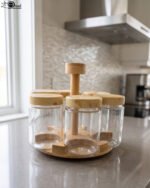 Six clear glass spice jars with smooth wooden lids arranged on a revolving wooden carousel stand on a kitchen countertop.