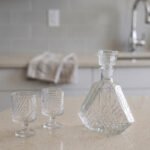 Crystal glass water bottle decanter with stopper alongside matching patterned stemmed glasses on a kitchen counter.