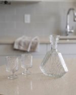 Crystal glass water bottle decanter with stopper alongside matching patterned stemmed glasses on a kitchen counter.