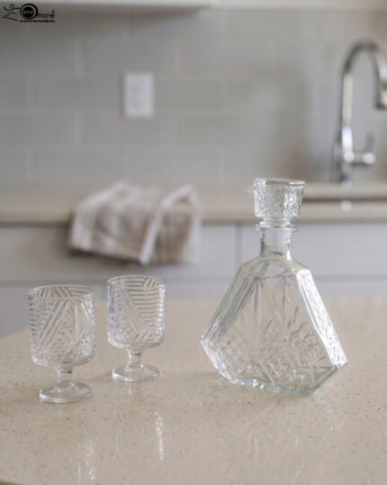 Crystal glass water bottle decanter with stopper alongside matching patterned stemmed glasses on a kitchen counter.