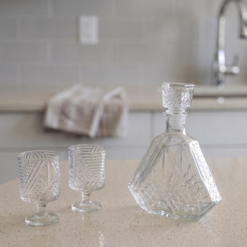 Crystal glass water bottle decanter with stopper alongside matching patterned stemmed glasses on a kitchen counter.