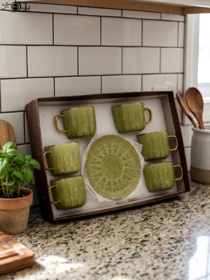 A boxed 6-person green ceramic cup and saucer set featuring textured patterns and gold handles and rims, displayed on a kitchen counter.