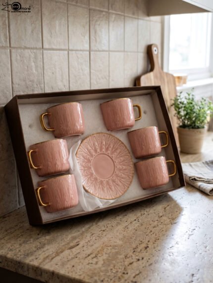 Pink ceramic cup and saucer 6-person set with gold trim inside a gift box on a kitchen counter.