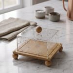 Modern square wooden cake dish with a clear acrylic lid featuring embedded silver leaf flakes and round wooden feet on a marble countertop.