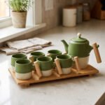 A modern green and cream ceramic tea set for 6 persons featuring wooden handles and a wooden tray on a kitchen counter.