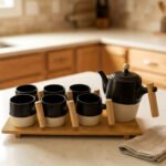 "A modern 6-person ceramic tea set displayed on a kitchen counter, including a teapot, six cups, and a bamboo serving tray, all featuring matte black and speckled finishes with wooden handles."