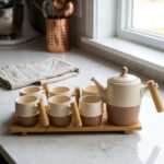 A complete modern tea set for 6 persons, featuring cream and mocha two-tone ceramic teapot and six matching mugs with bamboo handles, neatly organized on two bamboo serving trays on a kitchen counter.