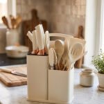 Off-white 19-piece silicone kitchen utensil and knife set with rose gold accents sitting in a dual caddy block on a countertop.