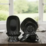 Glossy black 38-piece ceramic dinner set arranged on a rustic wooden dining table near a bright window.