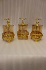 Three amber glass storage jars with gold deer lid handles on intricate gold filigree stands set on a kitchen marble countertop next to an espresso machine.