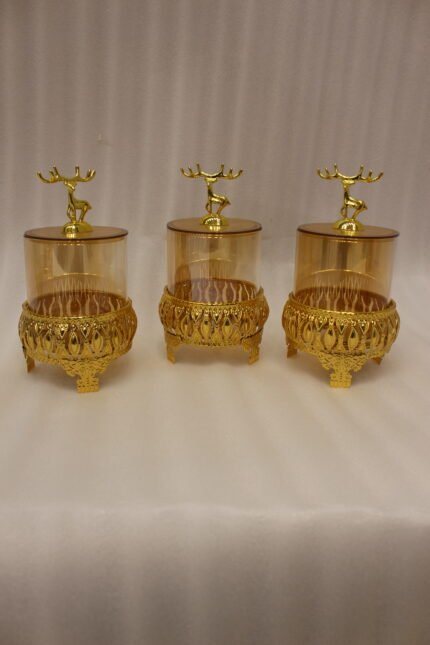 Three amber glass storage jars with gold deer lid handles on intricate gold filigree stands set on a kitchen marble countertop next to an espresso machine.