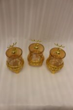 Three amber glass storage jars with gold deer lid handles on intricate gold filigree stands set on a kitchen marble countertop next to an espresso machine.