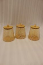 Three amber textured glass storage jars with natural wooden lids and crystal crystal-cut handles sitting on a marble kitchen countertop.