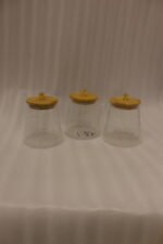 Three hammered glass kitchen storage jars with wooden wooden lids and crystal handles on a white marble countertop