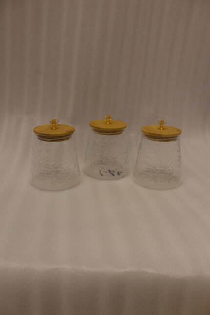 Three hammered glass kitchen storage jars with wooden wooden lids and crystal handles on a white marble countertop