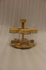 Six clear glass spice jars with smooth wooden lids arranged on a revolving wooden carousel stand on a kitchen countertop.