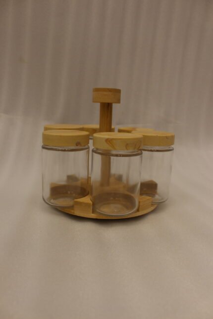 Six clear glass spice jars with smooth wooden lids arranged on a revolving wooden carousel stand on a kitchen countertop.