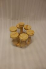 Six clear glass spice jars with smooth wooden lids arranged on a revolving wooden carousel stand on a kitchen countertop.