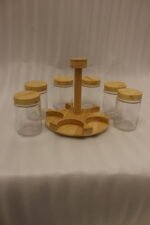 Six clear glass spice jars with smooth wooden lids arranged on a revolving wooden carousel stand on a kitchen countertop.