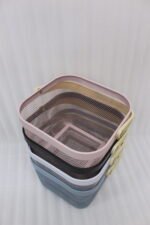 Stack of modern mesh wire storage baskets in pink, black, white, and blue with light wooden handles on a kitchen countertop.