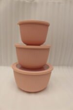Stack of three dusty pink ribbed plastic food storage bowls with clear lids on a white kitchen countertop.
