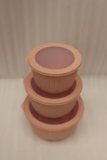 Stack of three dusty pink ribbed plastic food storage bowls with clear lids on a white kitchen countertop.