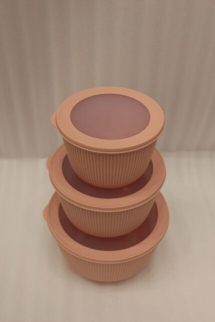 Stack of three dusty pink ribbed plastic food storage bowls with clear lids on a white kitchen countertop.