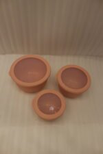 Stack of three dusty pink ribbed plastic food storage bowls with clear lids on a white kitchen countertop.