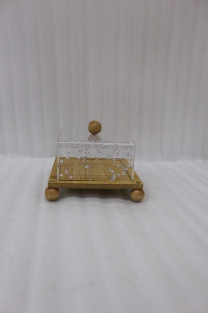 Modern square wooden cake dish with a clear acrylic lid featuring embedded silver leaf flakes and round wooden feet on a marble countertop.