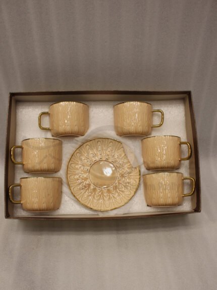 Elegant 6-person beige ceramic tea cup and saucer set with gold handles arranged inside a presentation gift box on a kitchen counter.