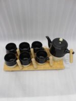 "A modern 6-person ceramic tea set displayed on a kitchen counter, including a teapot, six cups, and a bamboo serving tray, all featuring matte black and speckled finishes with wooden handles."