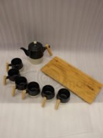 "A modern 6-person ceramic tea set displayed on a kitchen counter, including a teapot, six cups, and a bamboo serving tray, all featuring matte black and speckled finishes with wooden handles."