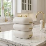 Three stacked off-white insulated hot pot casseroles with metallic gold handles sitting on a marble kitchen countertop.