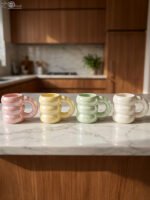 Four iridescent chubby ceramic mugs in pastel pink, yellow, mint green, and white displayed on a marble countertop.Four iridescent chubby ceramic mugs in pastel pink, yellow, mint green, and white displayed on a marble countertop.